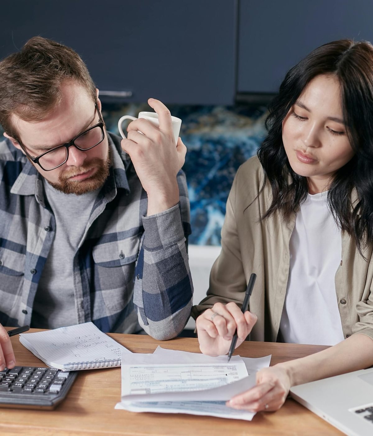 A couple reviewing household bills and budget using a calculator and laptop at their kitchen table.